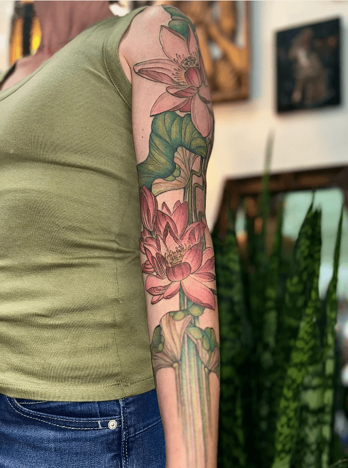 Person with a colorful floral tattoo on their arm, wearing a green sleeveless top and blue jeans, indoors.