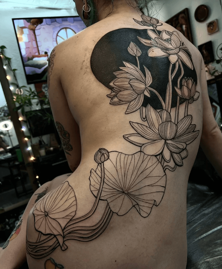 Back tattoo of flowers and leaves with bold outlines on person's back and shoulder, in a room with plants and decor.