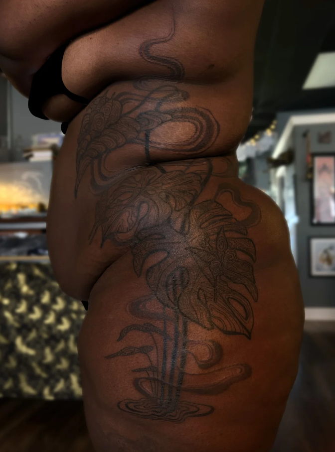 Side view of a person with a large tattoo of a leaf and floral design on their torso and hip, in a room with framed pictures.