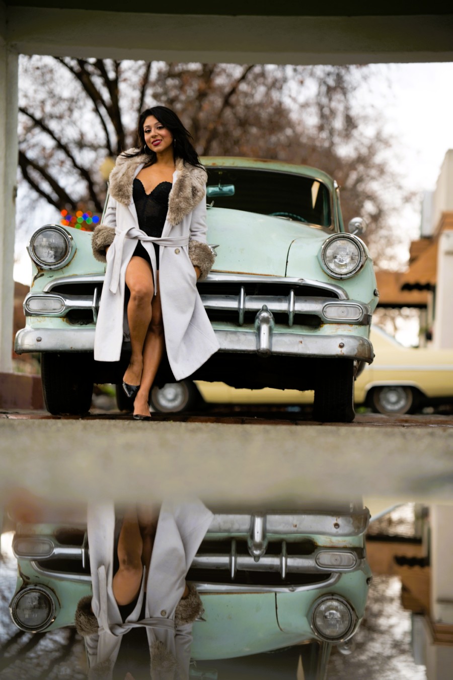 Woman in black dress and coat standing in front of vintage car, reflection visible on wet ground.