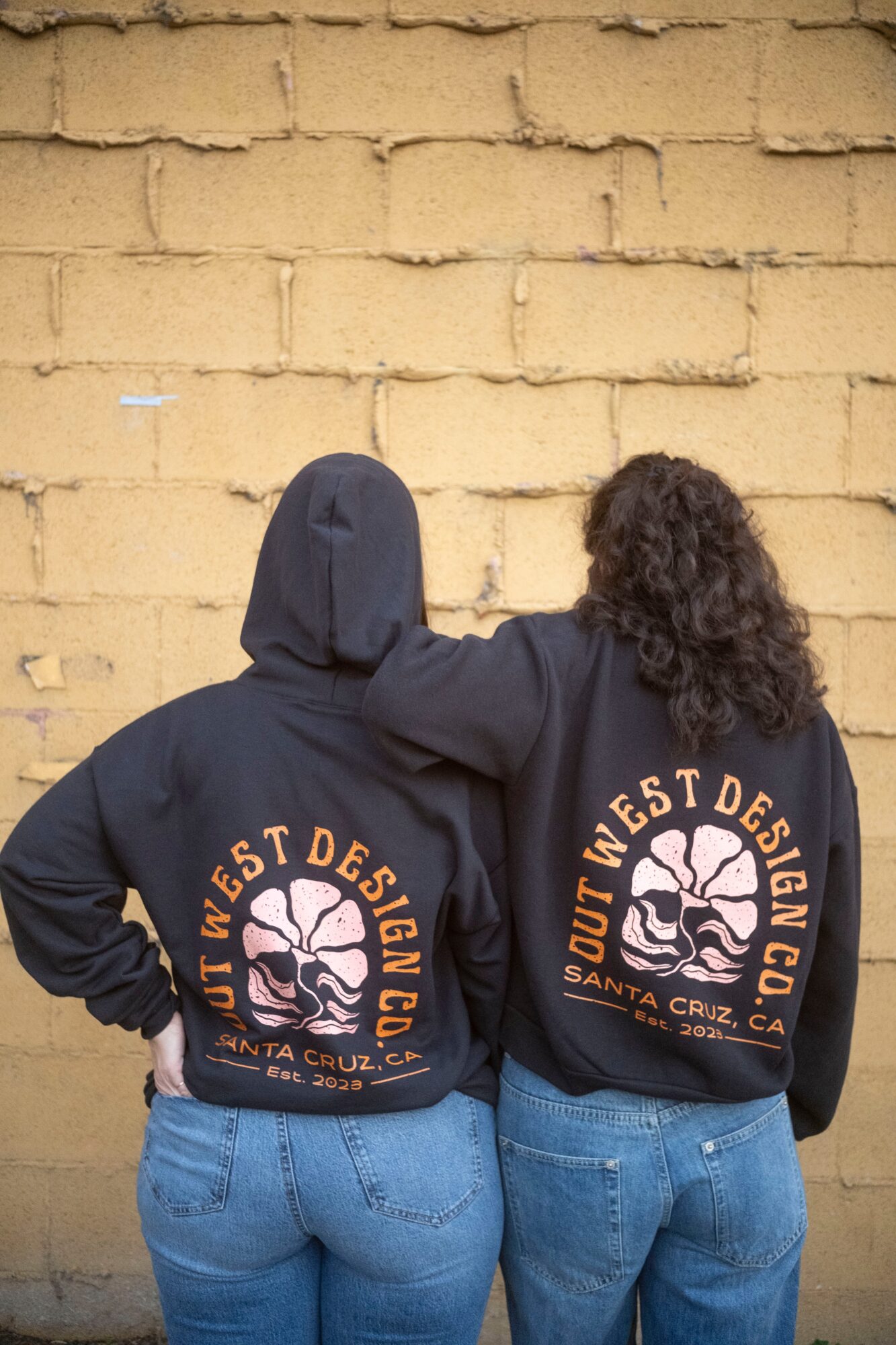 Two people wearing black hoodies with a logo on the back, standing against a yellow brick wall, facing away.
