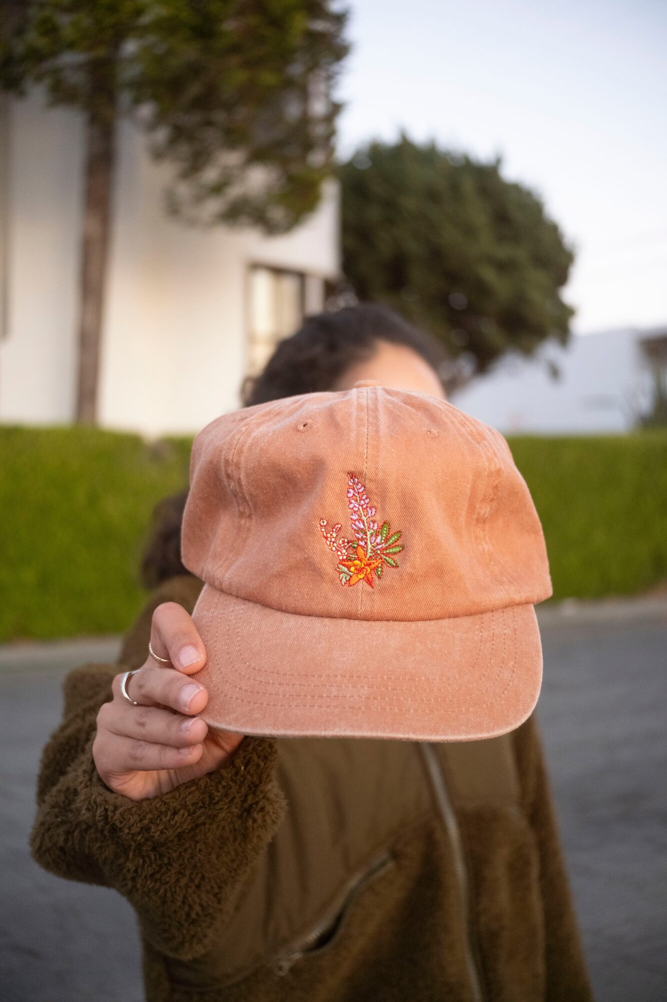 Person holding a peach-colored cap with floral embroidery in front of face outdoors.