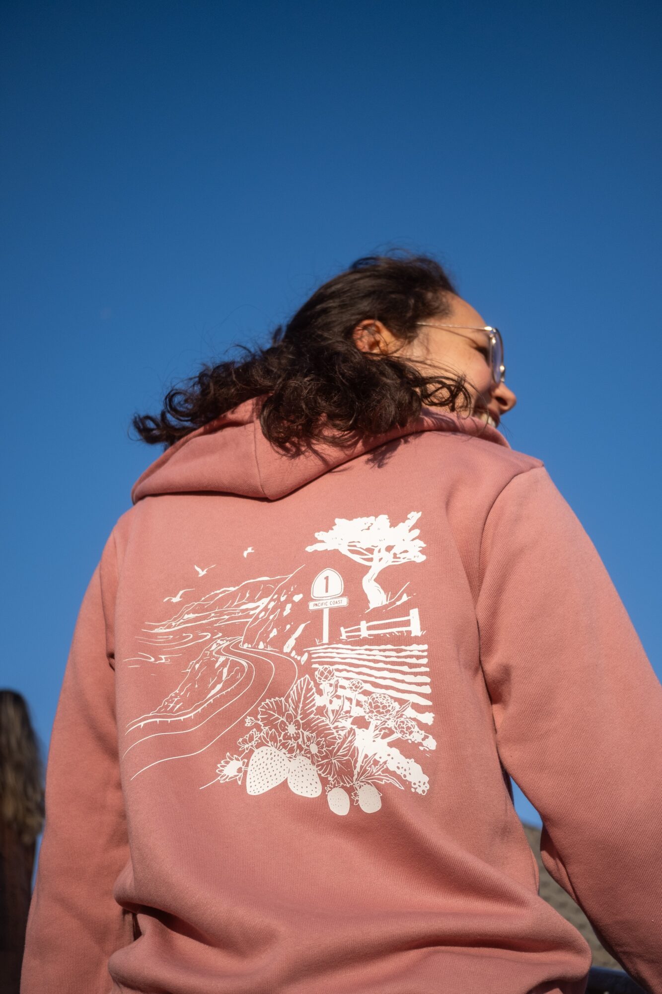 Person with curly hair wearing a pink hoodie with a white graphic design on the back, facing away from the camera, against a clear blue sky.