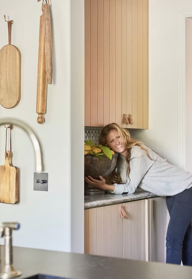 Woman leaning on kitchen counter holding a bowl, smiling, with wooden cabinets and hanging kitchen utensils visible.
