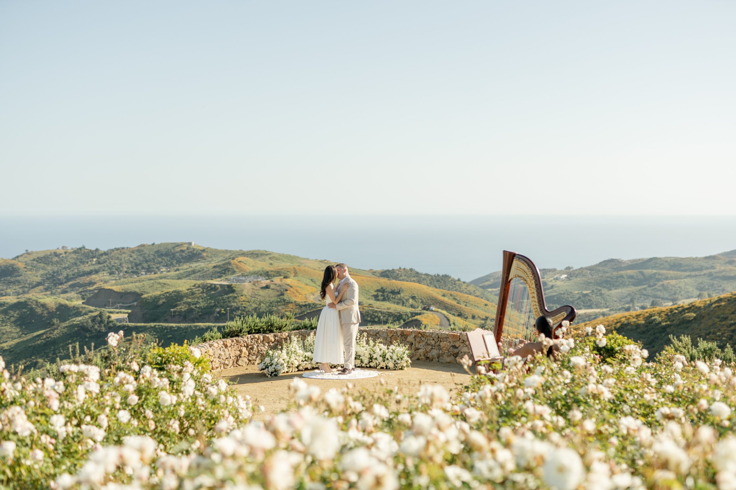 A woman in a white dress stands outdoors near a mirror, with a landscape of hills and the sea in the background.