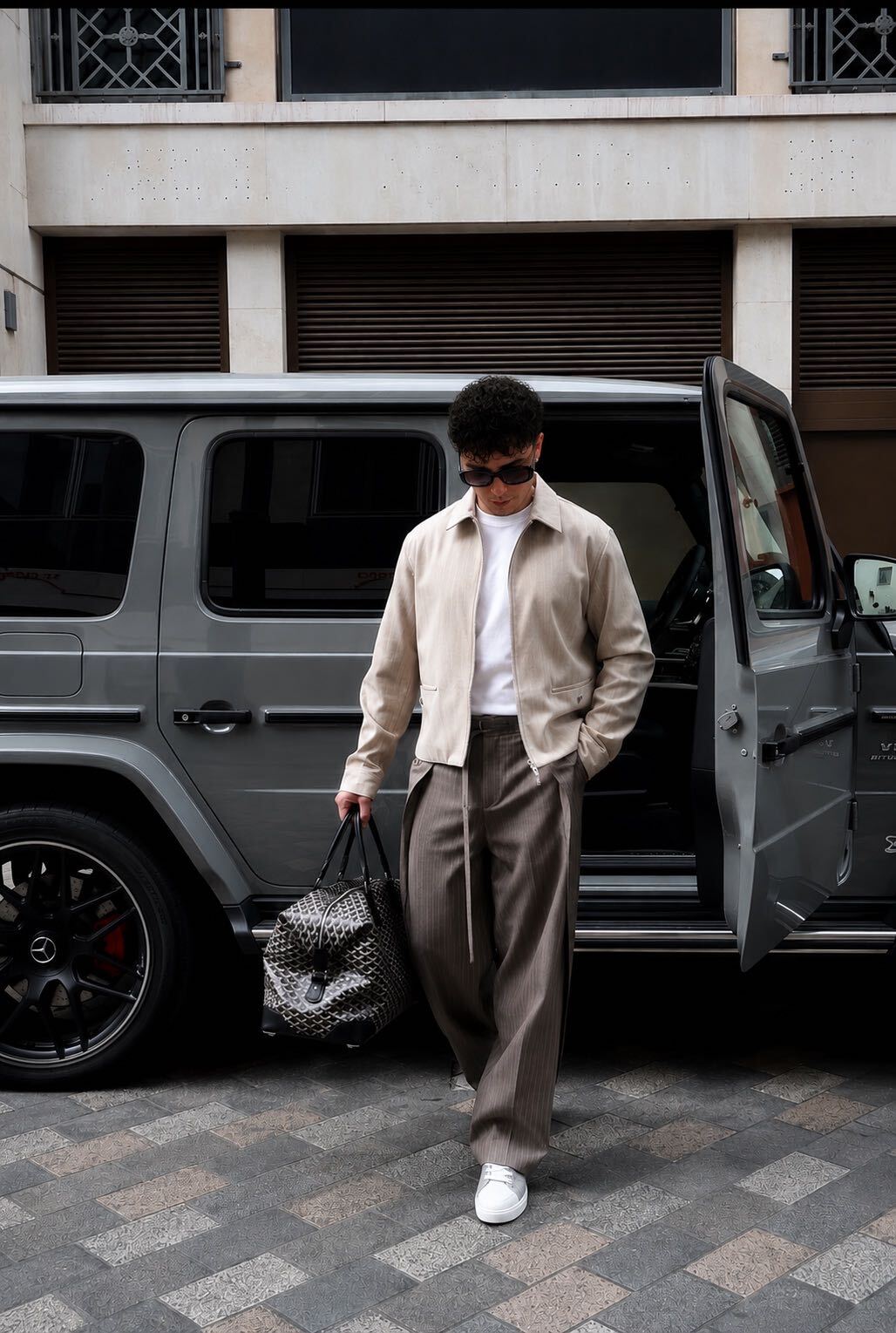 Person with sunglasses, beige jacket, white shirt, gray pants, white shoes, walking away from a black vehicle, carrying a patterned bag.
