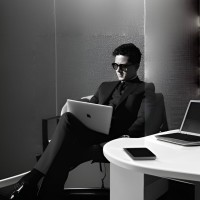 Person sitting on a chair using a laptop, with a table holding a smartphone and another laptop nearby.
