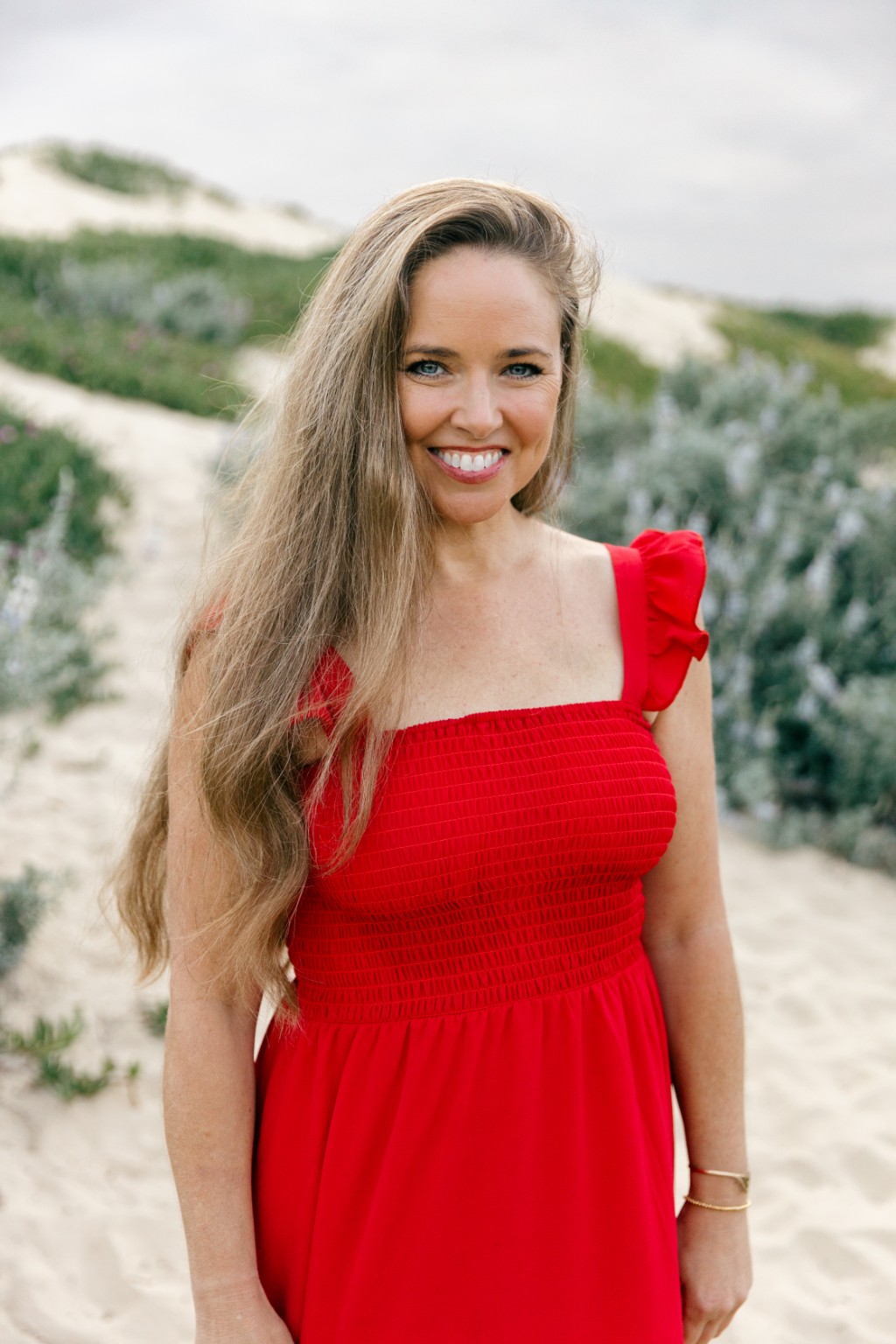 Smiling woman with long wavy hair in a red dress outdoors, sandy path and greenery in background.