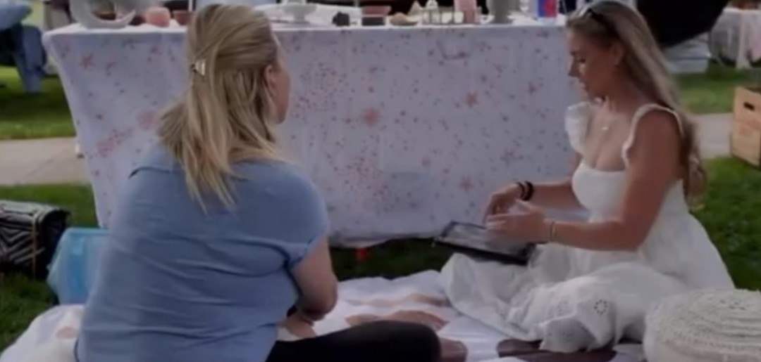 Two women sitting on the ground outdoors, one in a blue shirt and the other in a white dress, talking.