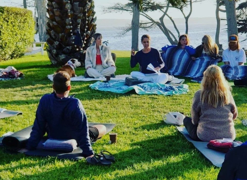 Group of people sitting on mats and blankets on grass near trees and water, participating in outdoor activity.