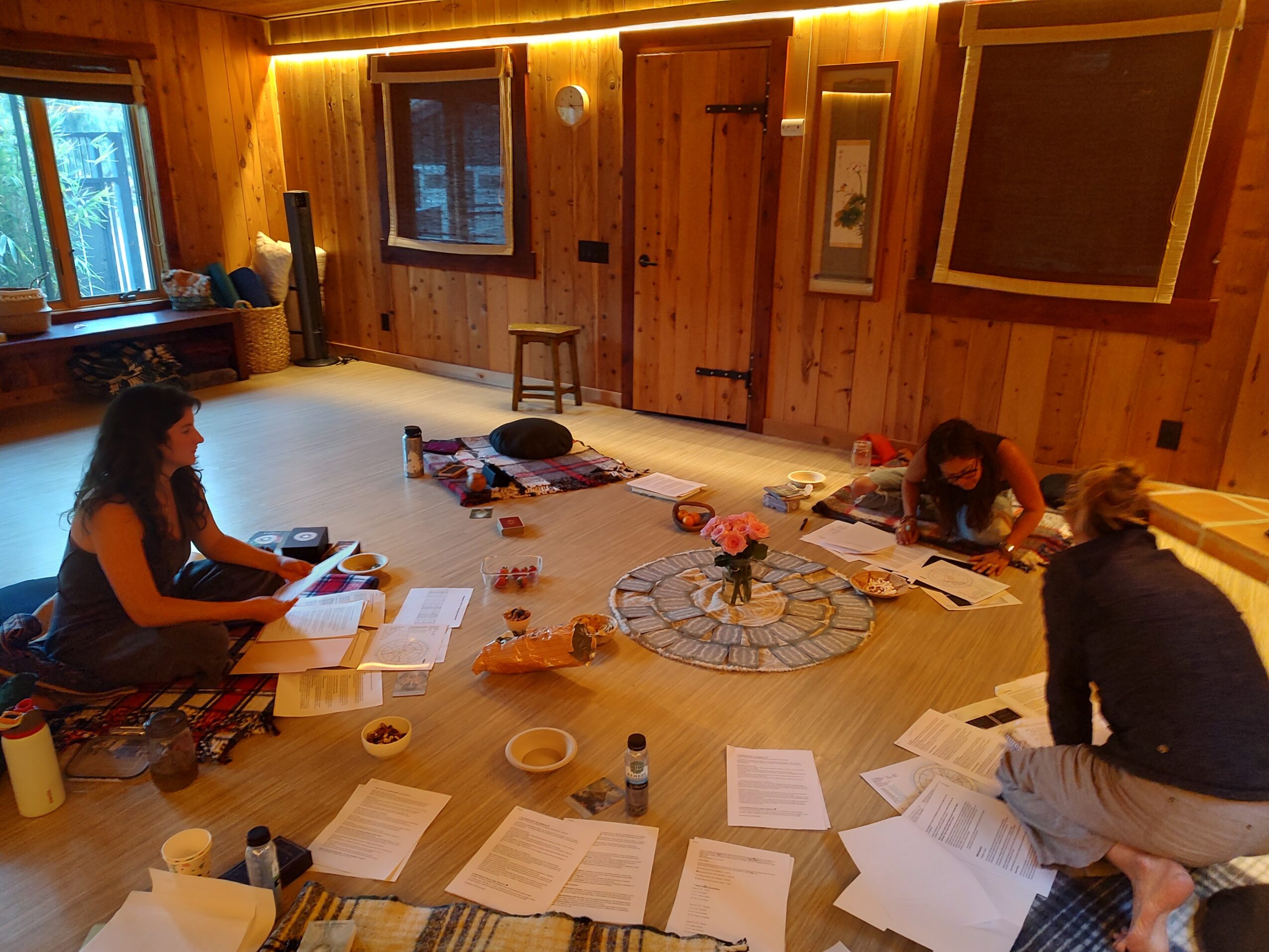 Three people sitting on the floor with papers, books, and snacks in a wooden room with windows and warm lighting.