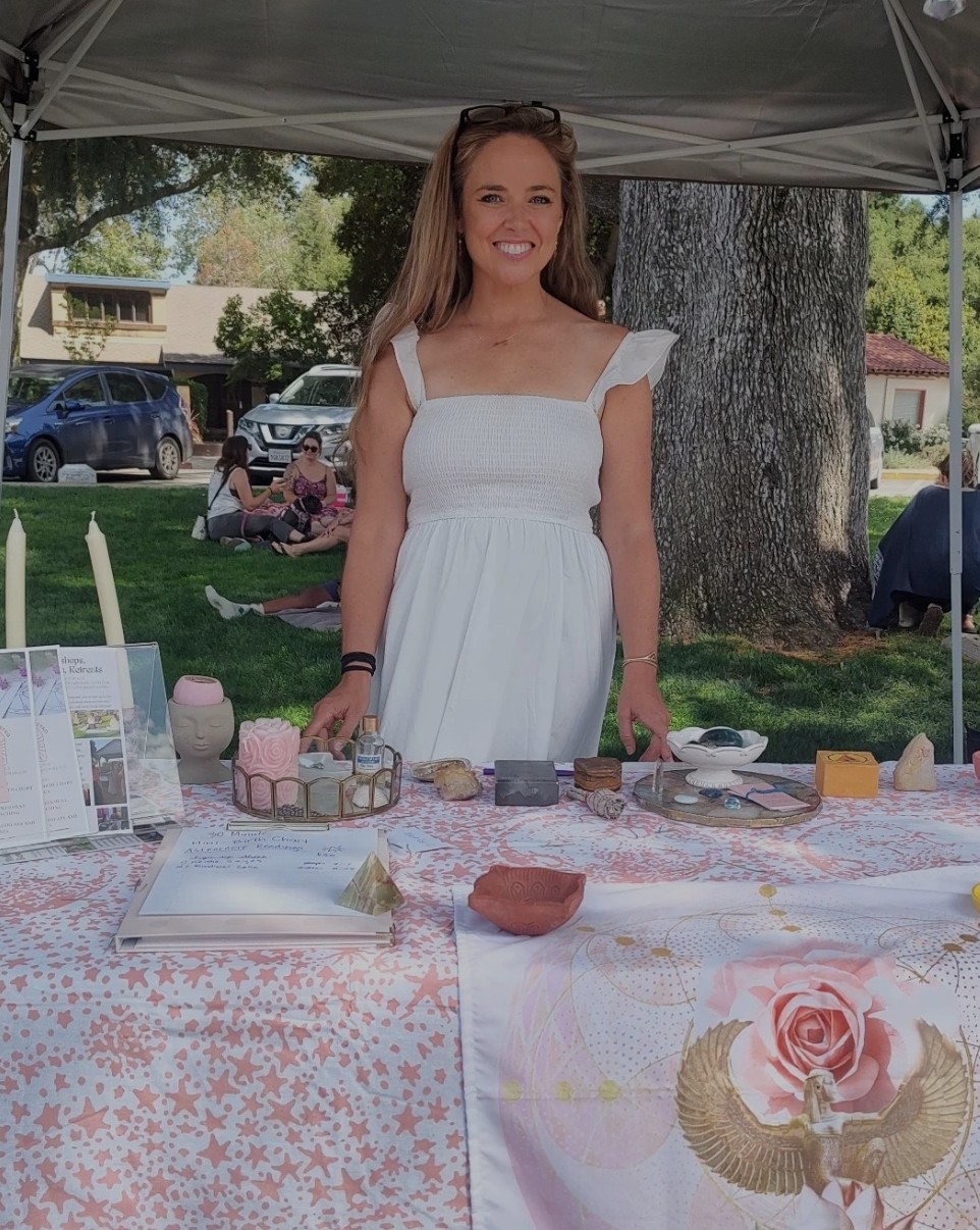 Young woman in white dress standing behind a table outdoors, smiling, with trees and cars in background.