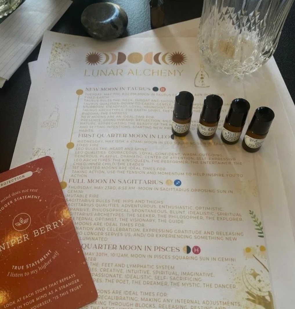 Four small bottles of essential oils are placed on a paper with lunar alchemy symbols and text, next to a glass and a red card.