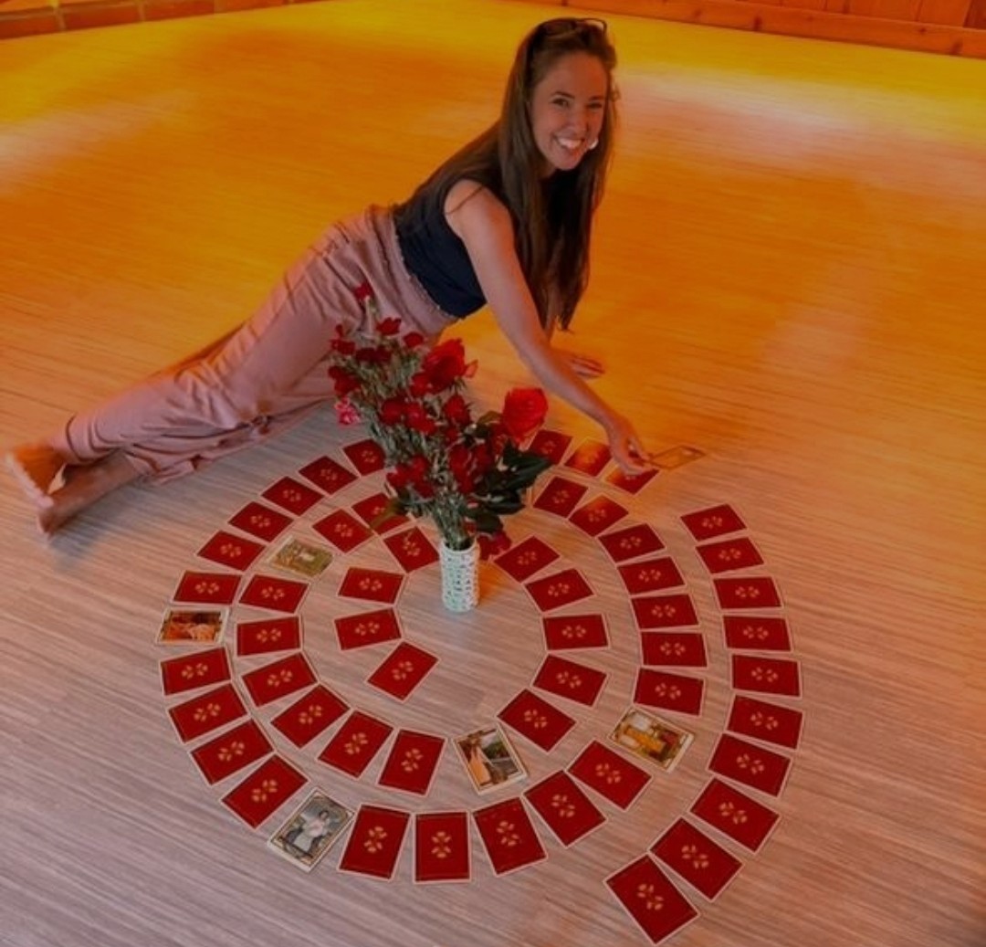 Girl lying on the floor with a card circle, flowers, and a book, smiling at the camera in a warm room.