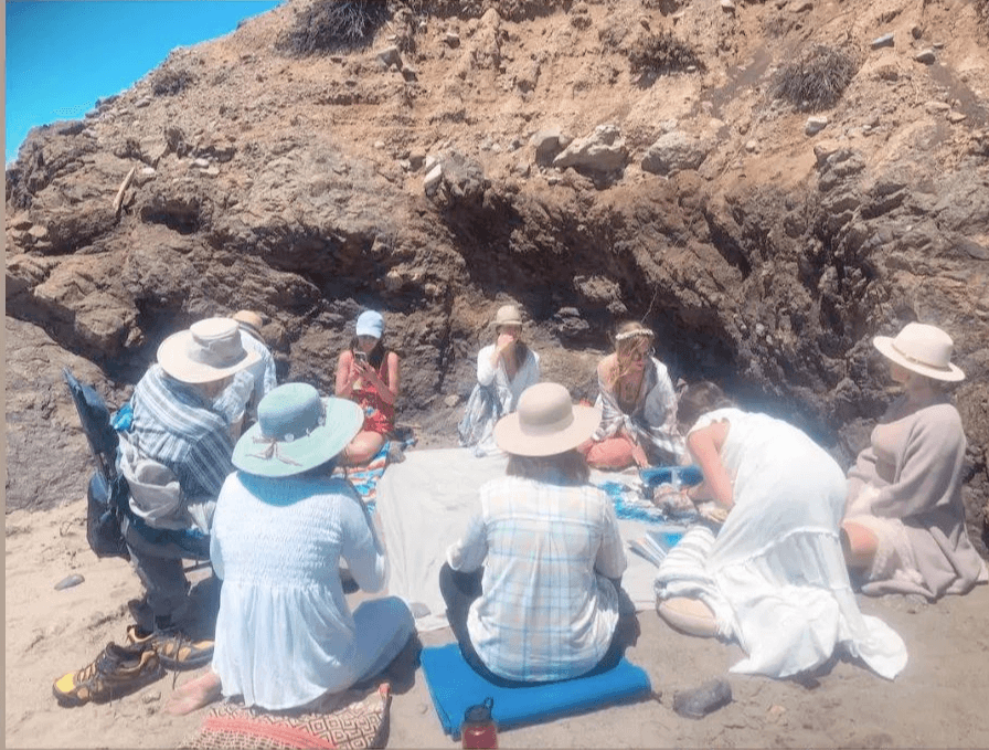 Group of people sitting on mats and rocks on a beach near a large rocky cliff, some wearing hats.
