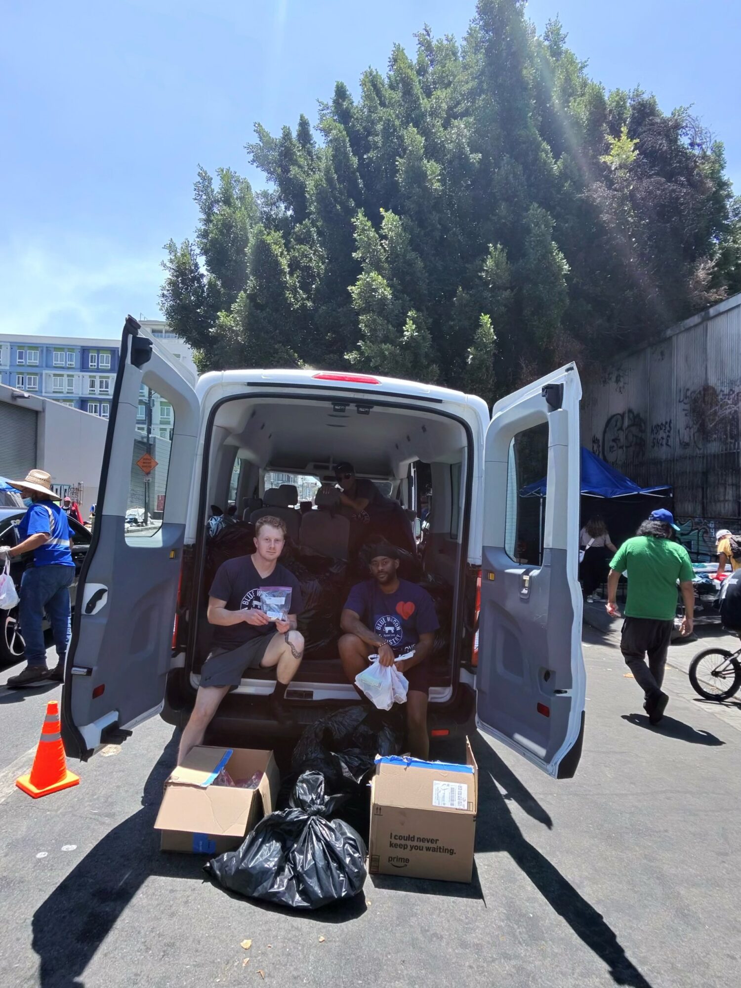Two people sitting inside a van with open doors, surrounded by boxes and bags, outdoors on a sunny day.