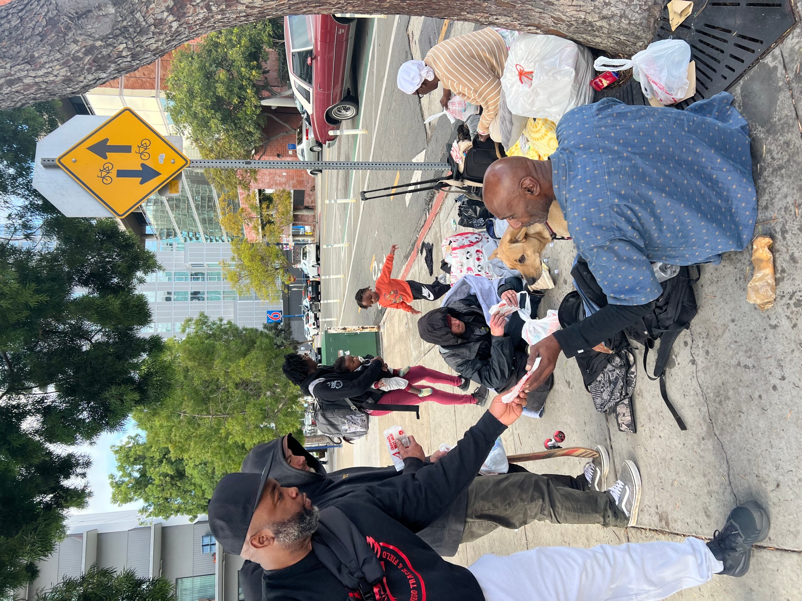 Person sitting on the sidewalk reaching out to help another person, with others nearby and a street scene in background.