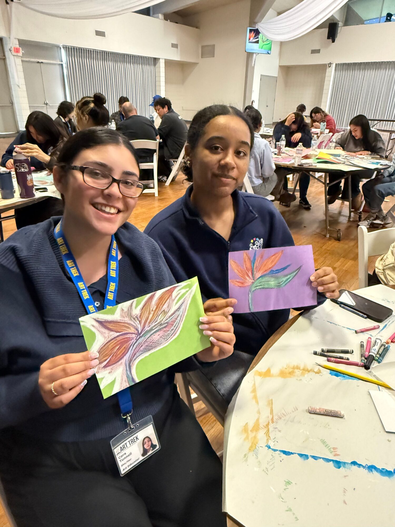 Two women sitting at a table holding colorful drawings, smiling, in a busy classroom or workshop setting.