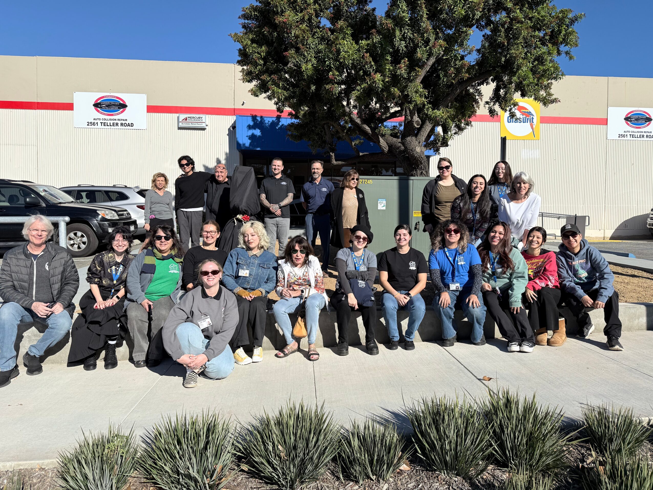 Group of people posing outdoors in front of a building with trees and parked cars, some sitting and some standing.