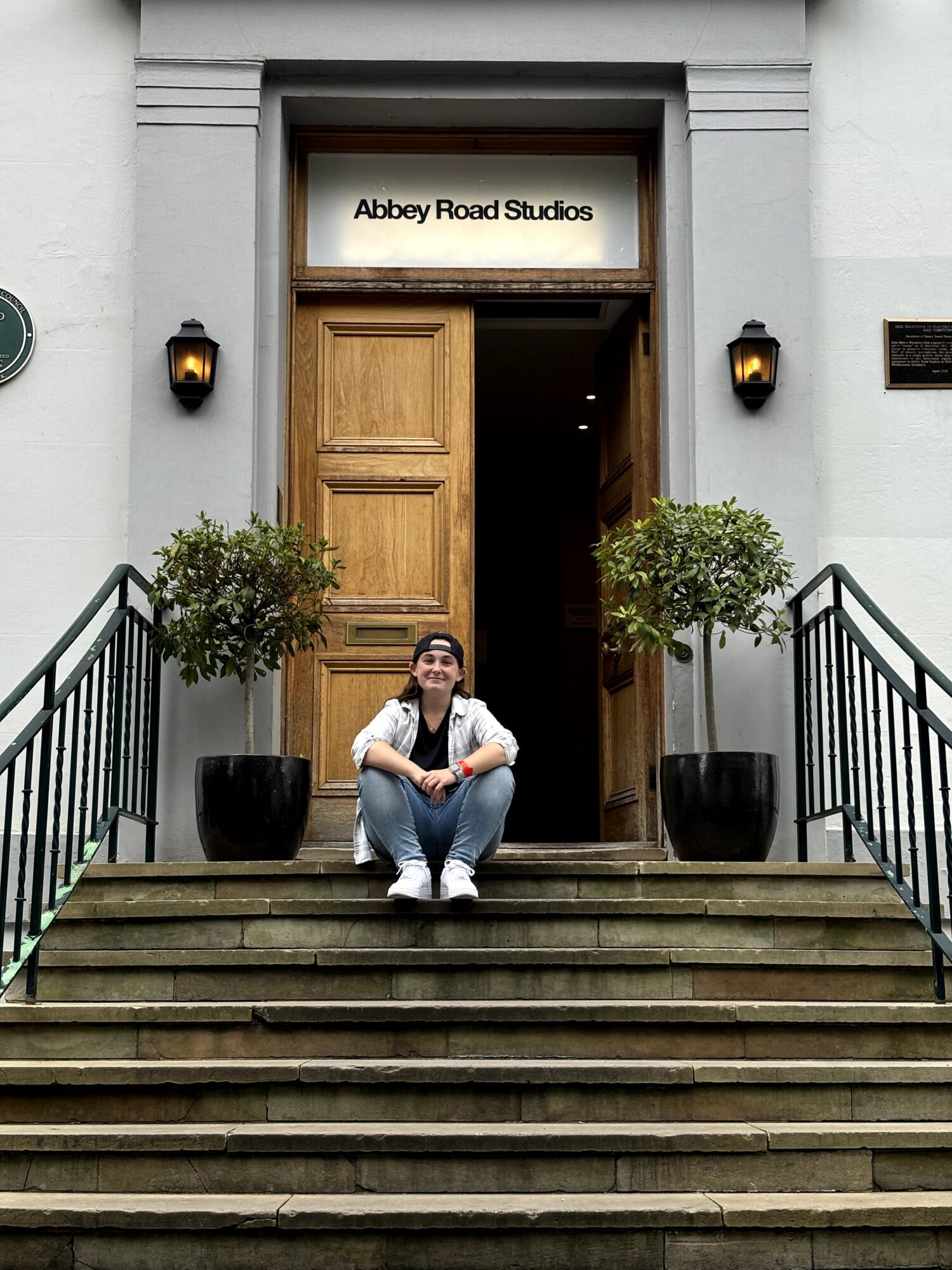 Person sitting on stairs outside building with open wooden door, two potted plants, and sign reading 'Abbey Road Studios'.