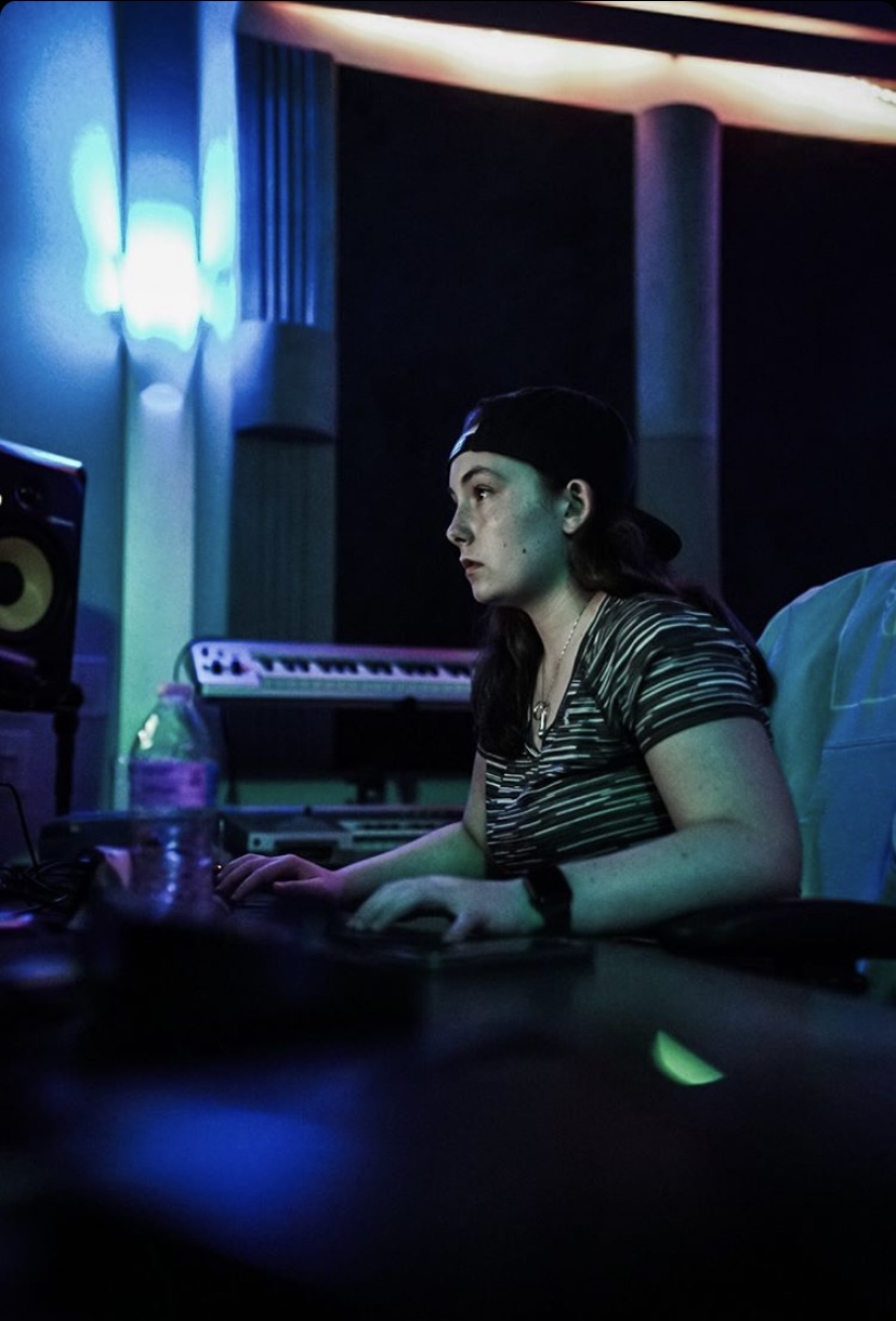Woman with dark hair in a striped shirt working at a music studio with blue lighting, keyboard, and sound equipment
