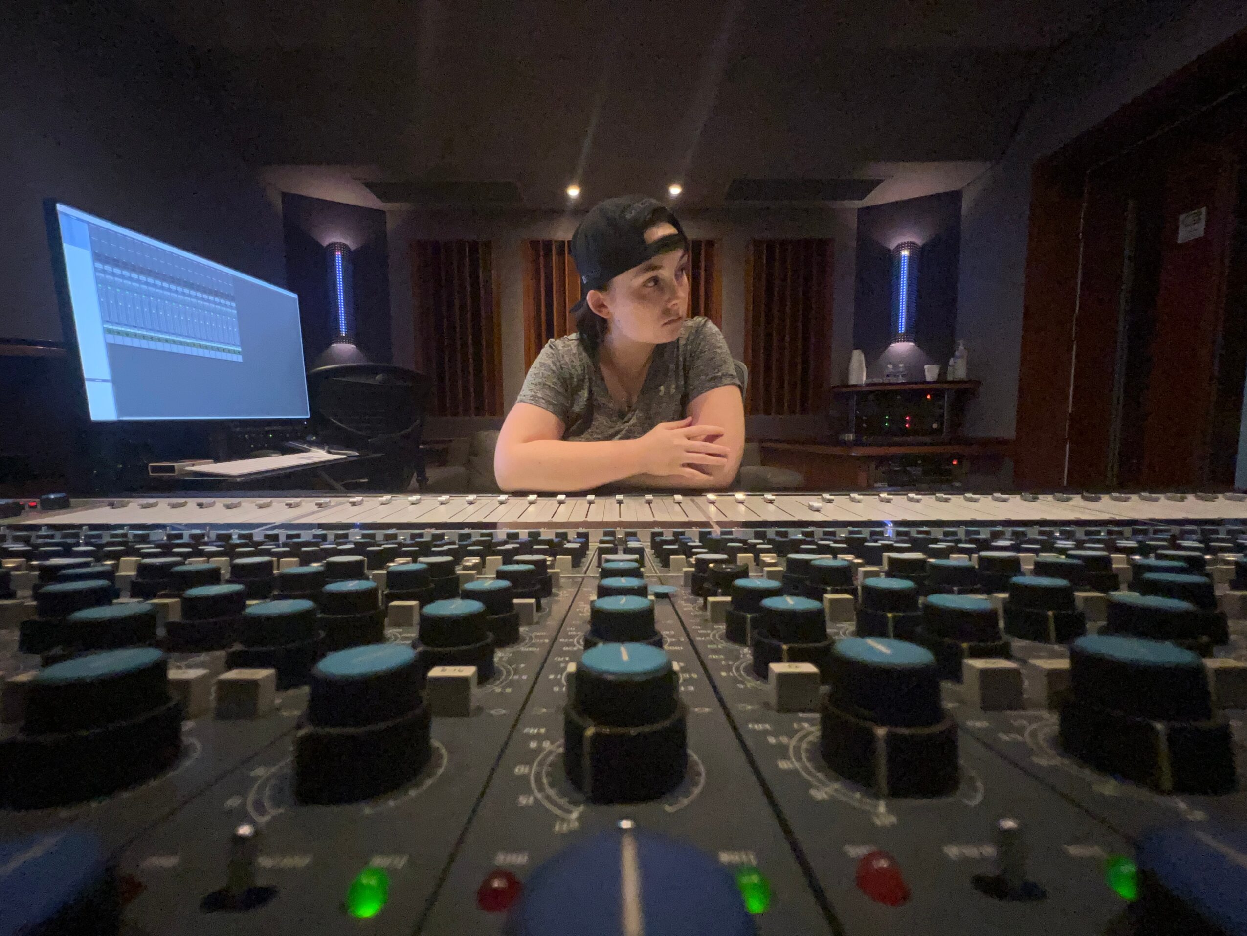 Person sitting at a mixing console in a recording studio, with a computer screen in the background.