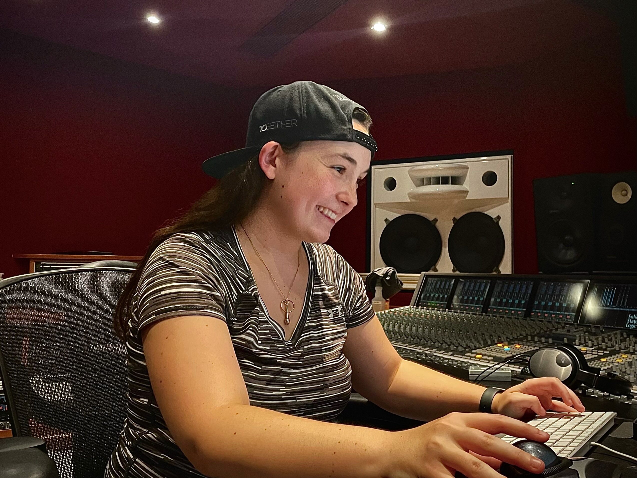 Young woman with long hair, wearing a striped shirt and backwards cap, working at a music mixing console in a studio.