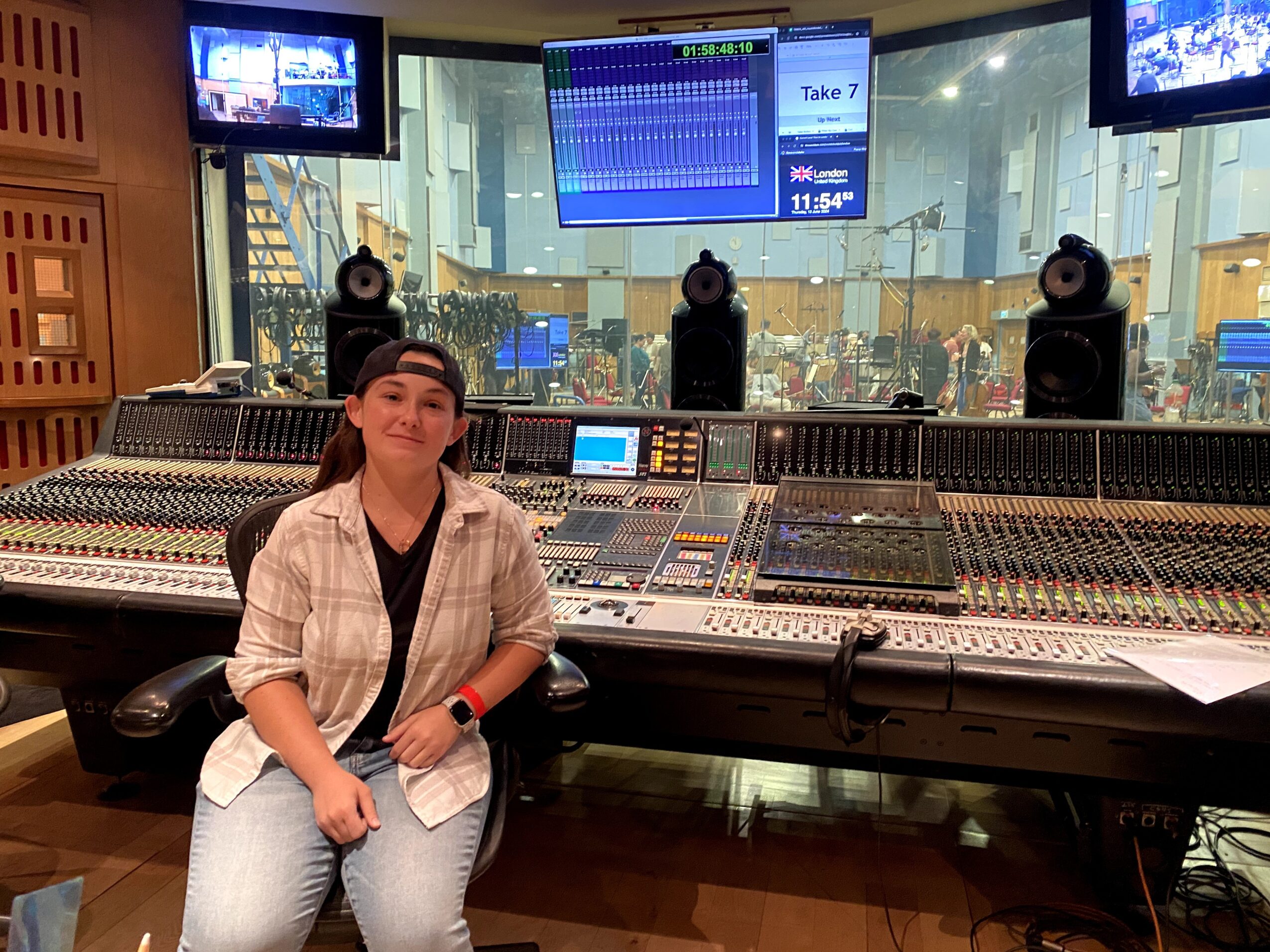 Person sitting in front of a large audio mixing console in a recording studio, with screens and speakers behind.