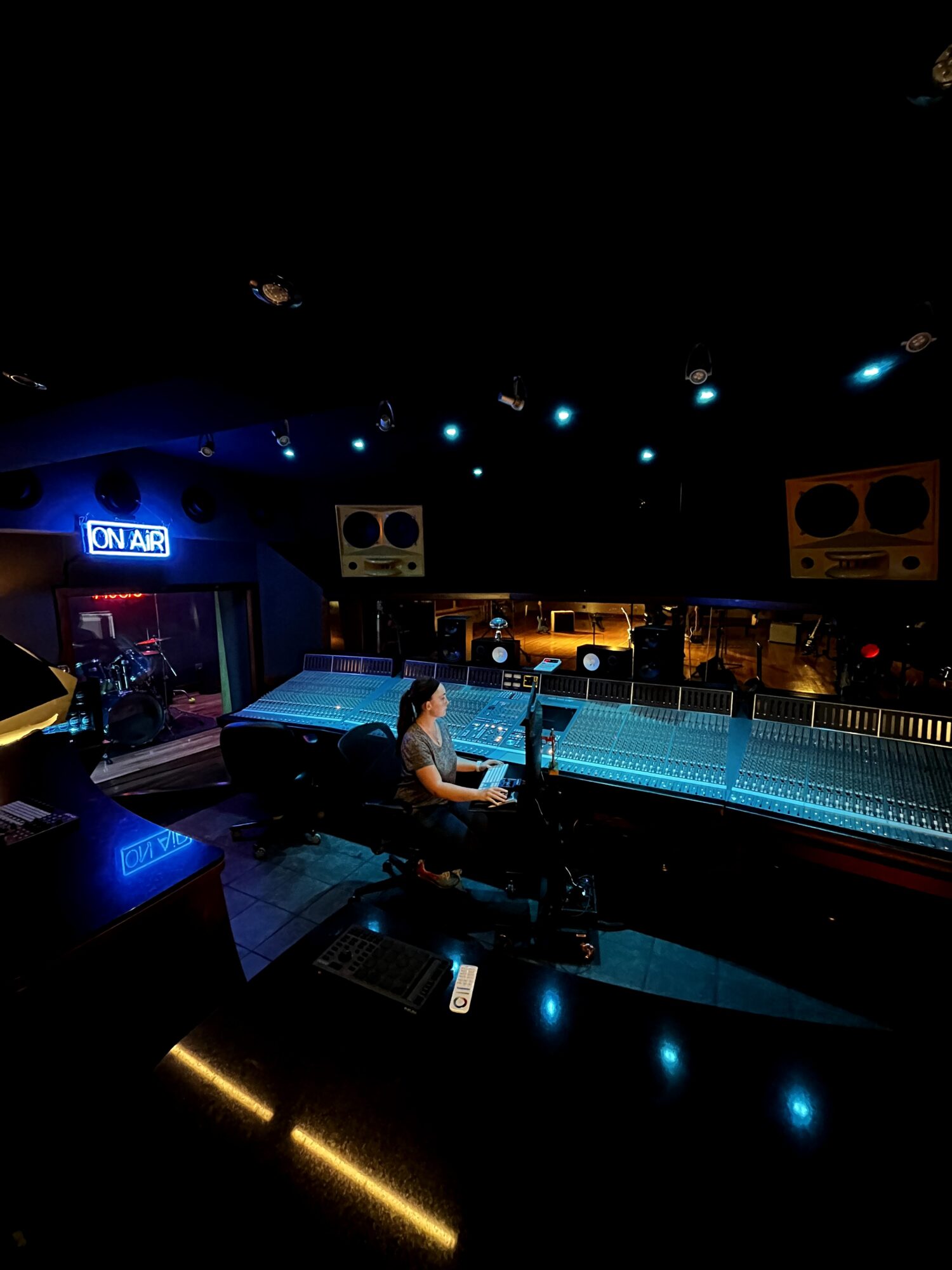 Person working at a sound mixing console in a dark recording studio with equipment and a window to another room.