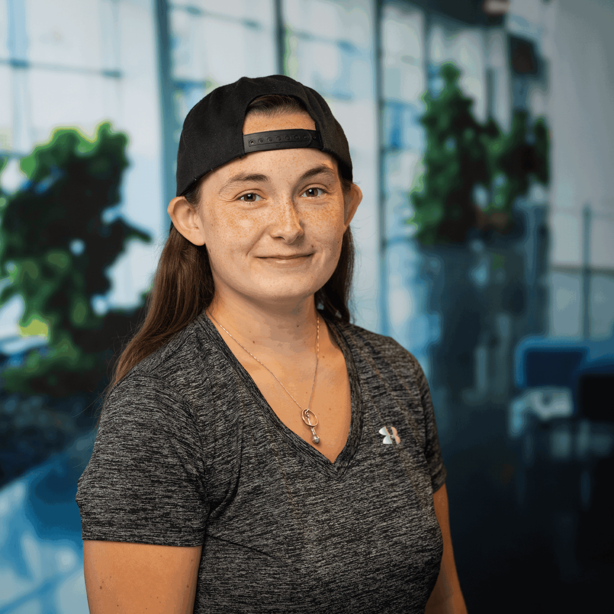 Young woman with long hair, wearing a black cap backwards and a dark gray shirt, smiling indoors with large windows and plants.