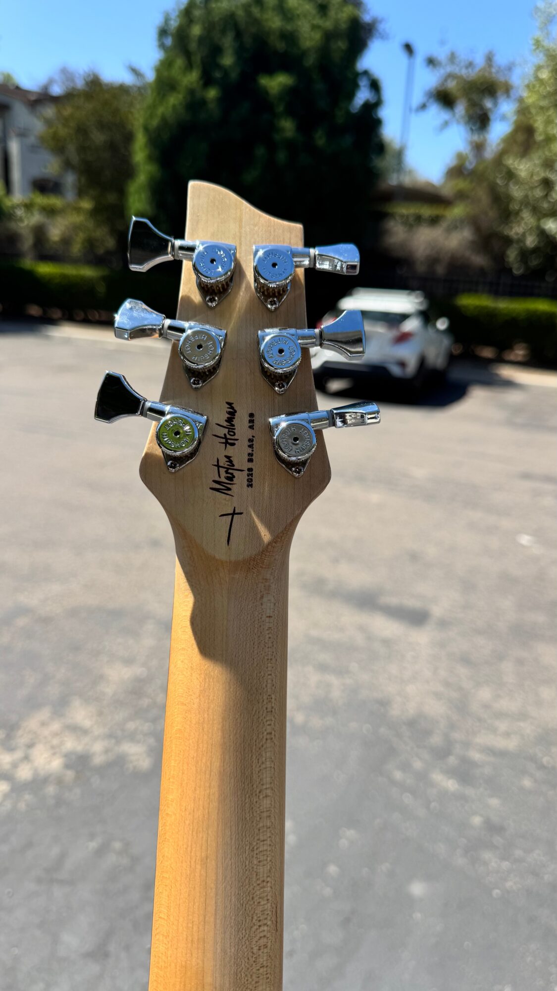 Guitar headstock with six tuning pegs, outdoors with trees and cars in background.