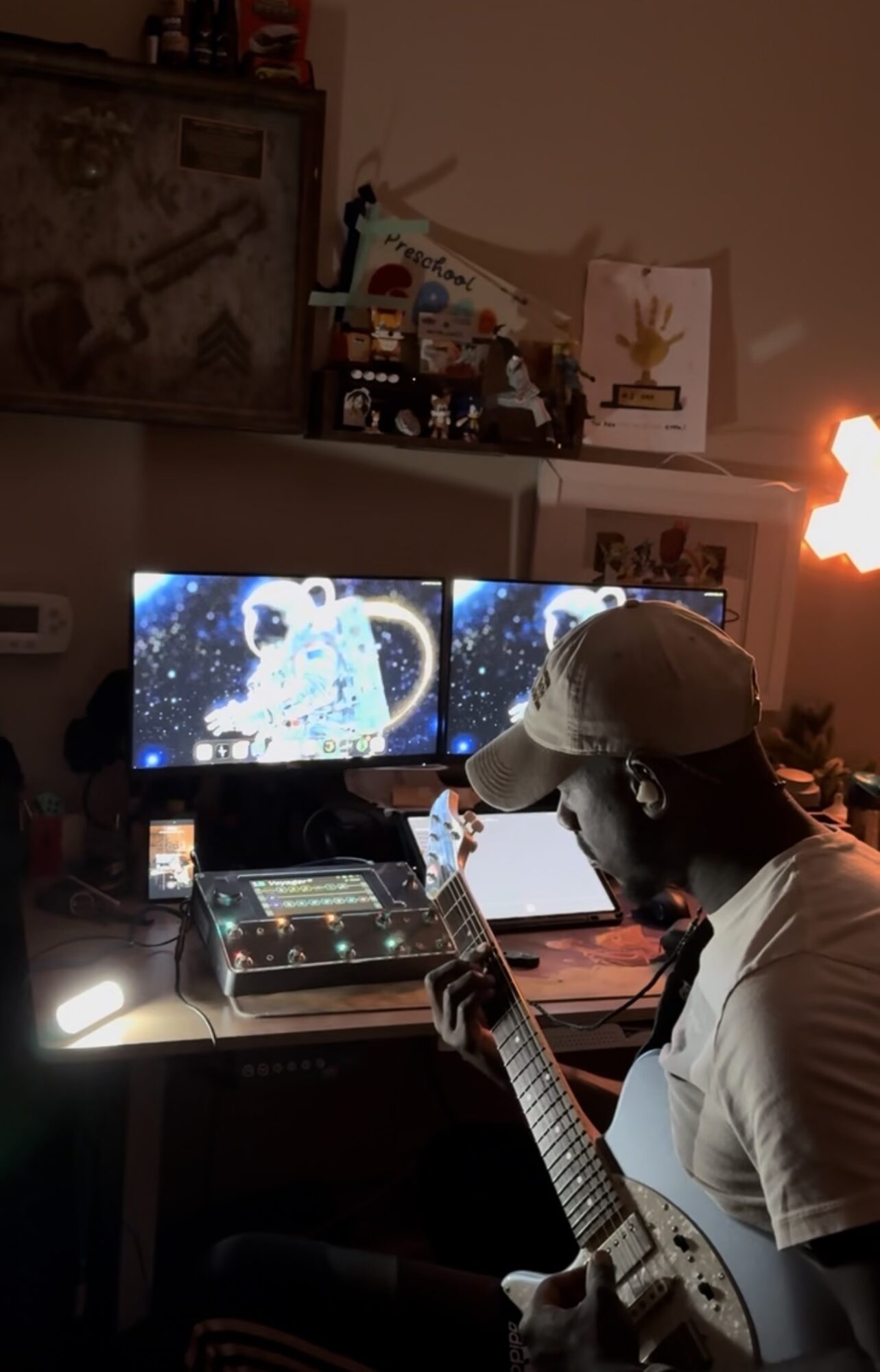 Person wearing a cap playing guitar in a room with two computer monitors displaying space images, and a lit candle or lamp on the right. Word count: 27