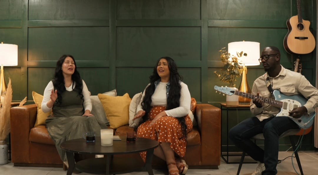 Three people sitting on a sofa in a room with green walls, a guitar hanging on the wall, and a lamp, singing and playing guitar.