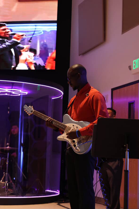 Person playing electric guitar in a dimly lit room with purple lighting, standing near a transparent cylindrical barrier.