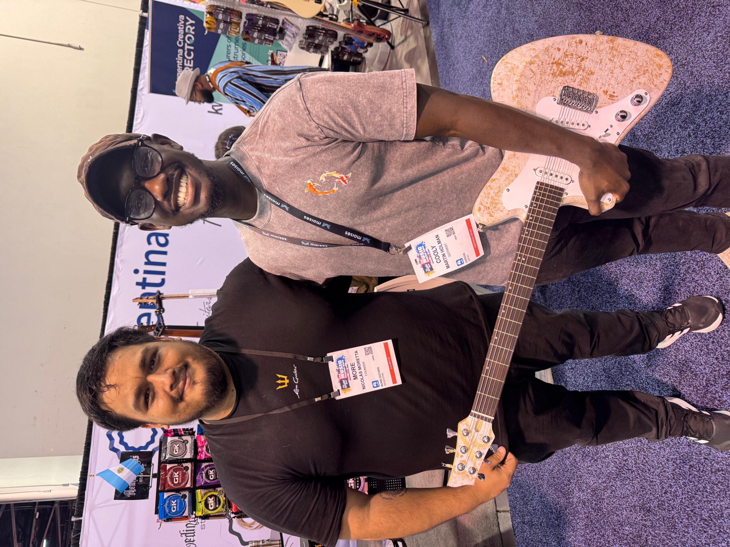 Two men standing at an indoor event, one holding a guitar, with booths and banners in the background.