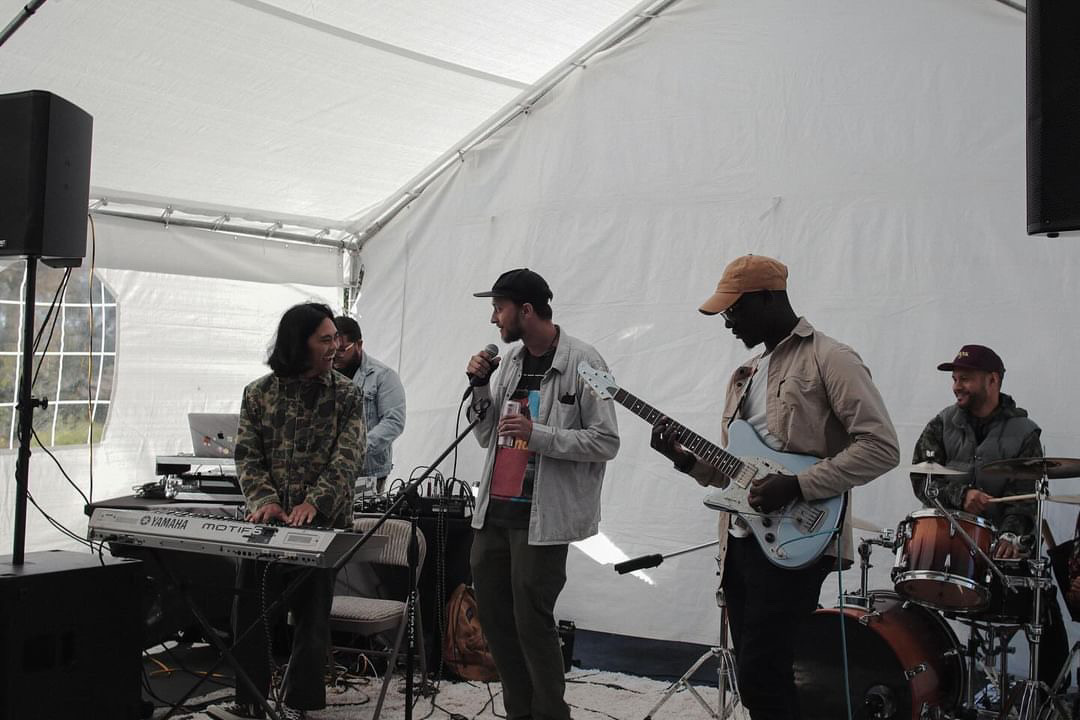 Four musicians perform inside a tent, with one playing keyboard, one singing, one playing guitar, and one on drums.