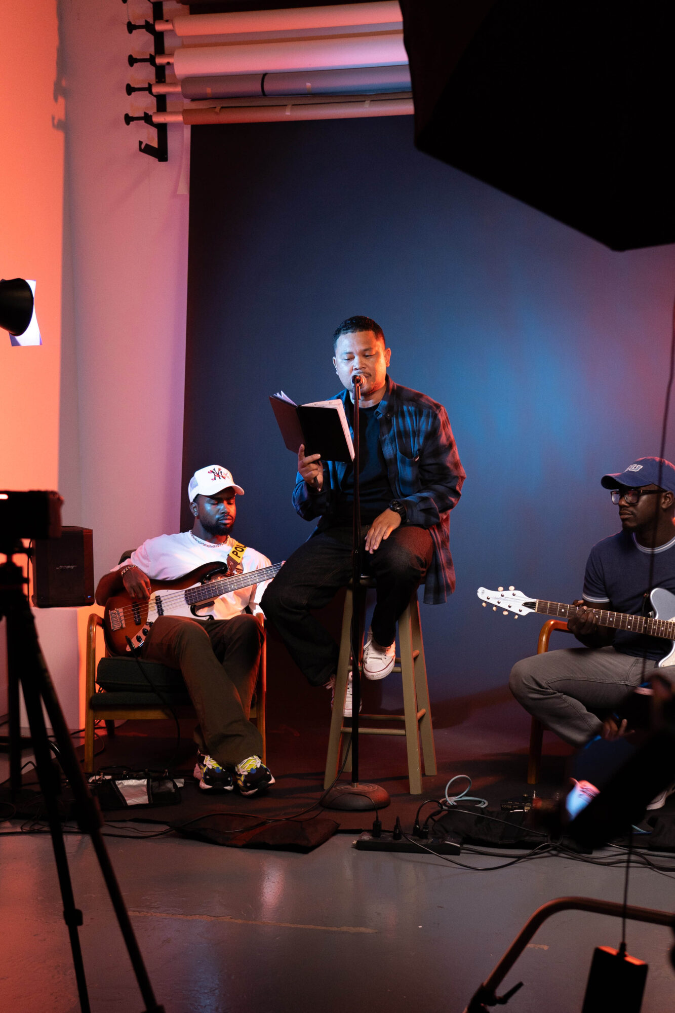 Person singing into a microphone with two guitar players on stage, one wearing a cap and glasses, in a performance setting.