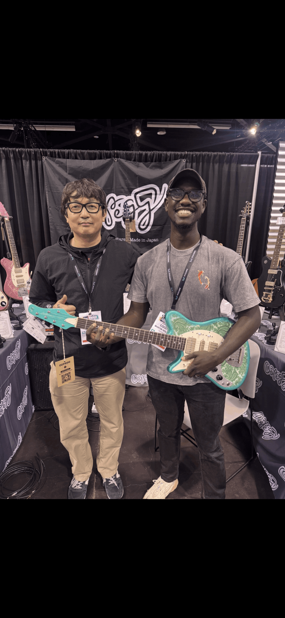 Two people standing with guitars in a music store, smiling, black curtain background, one with glasses, one with a beard.