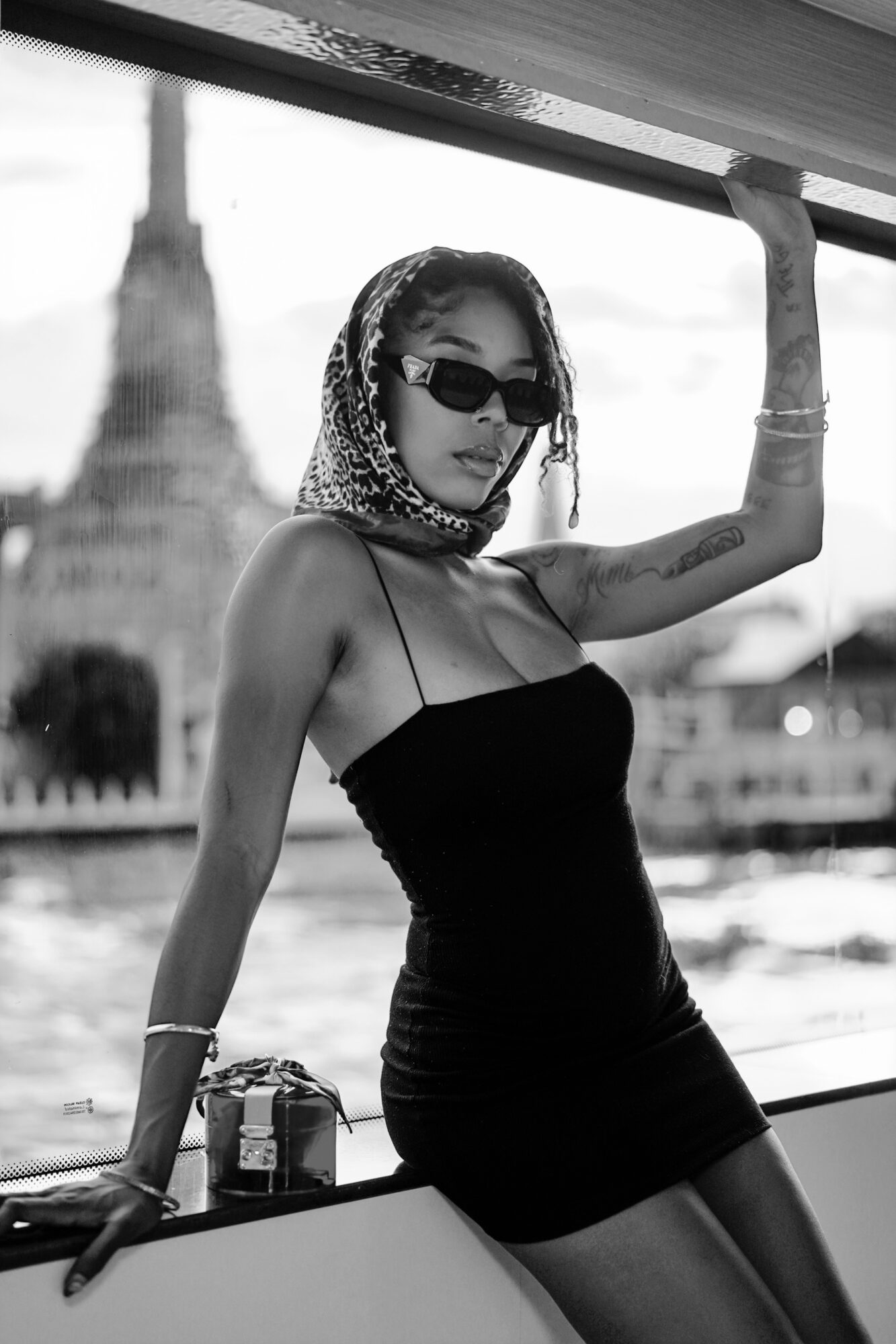 Woman in sunglasses and a headscarf leans against a boat railing outdoors, wearing a strapless black dress, with water and trees in background.