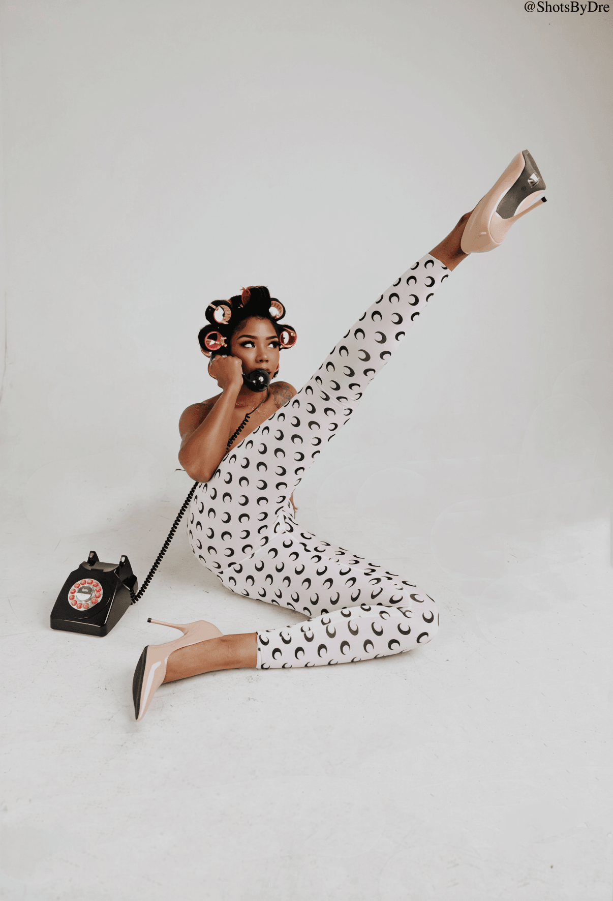 Young girl with hair rollers, wearing patterned jumpsuit, sitting on floor with one leg raised, holding a vintage telephone, against a plain background.
