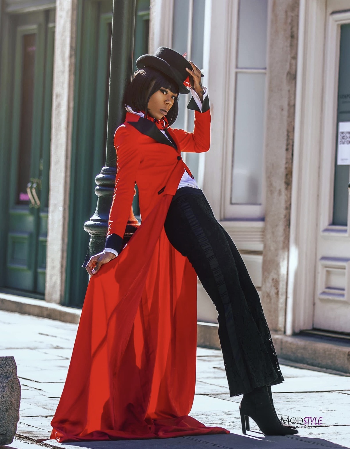 Woman in red coat and black pants poses on city street, holding hat, with buildings in background.