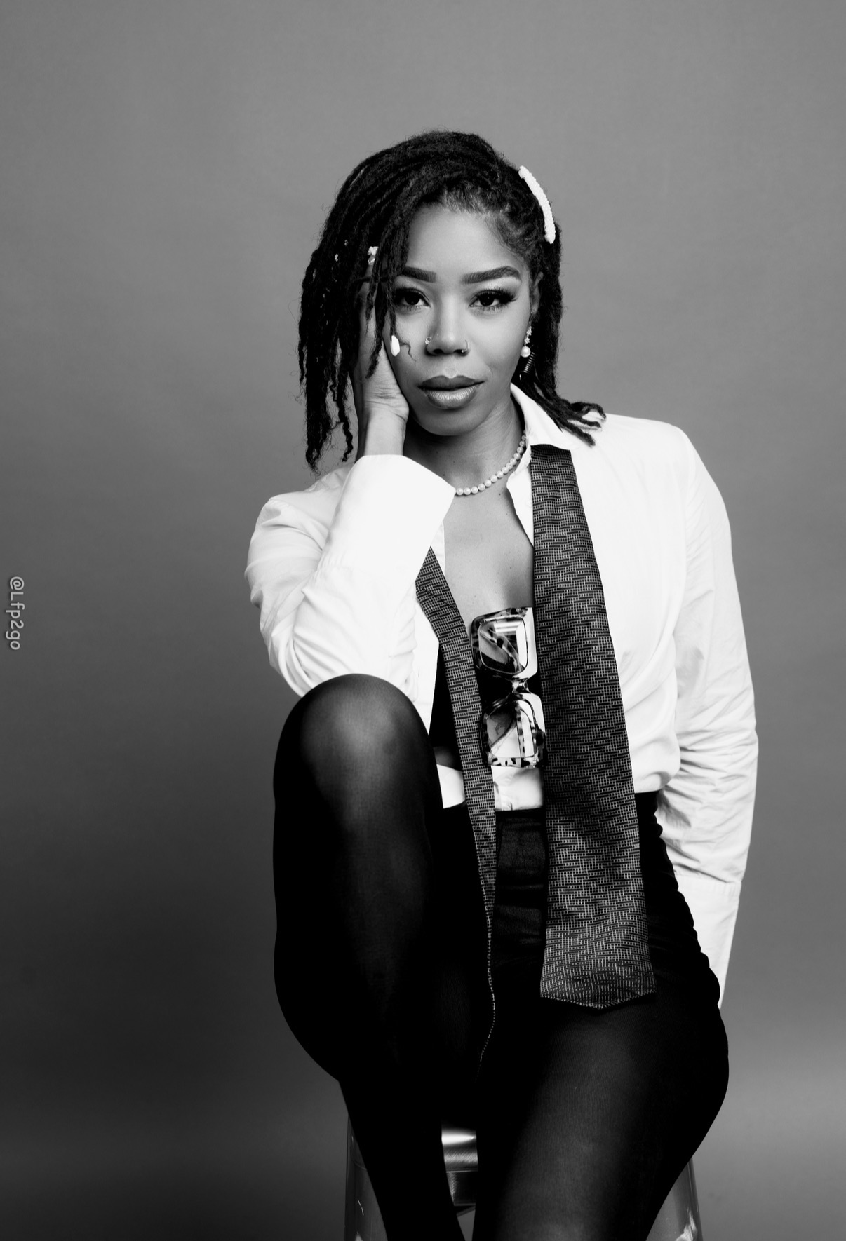 Portrait of a woman with dreadlocks, wearing a white blazer and dark pants, sitting with one knee raised, looking at the camera.