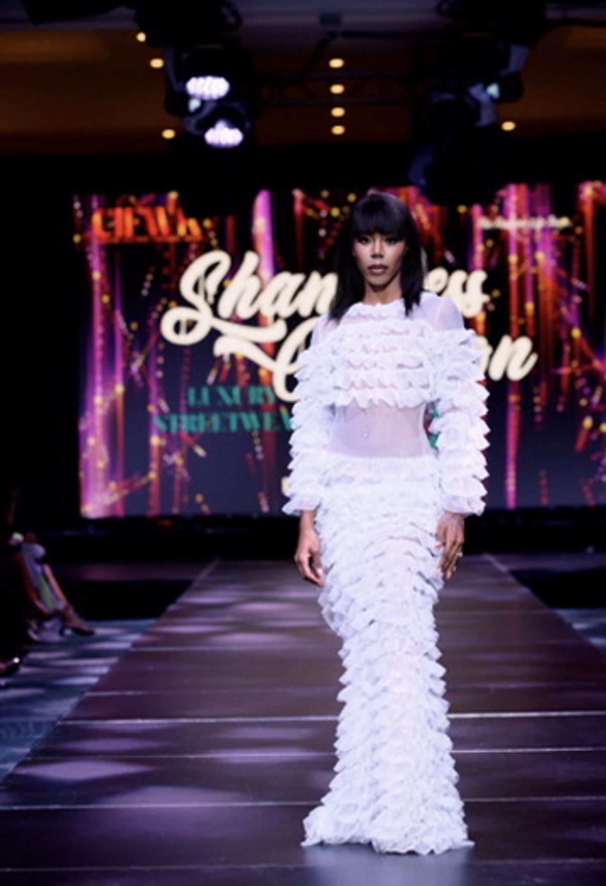 Female model walking on runway in white textured dress with fringe details, stage background with colorful lights and text