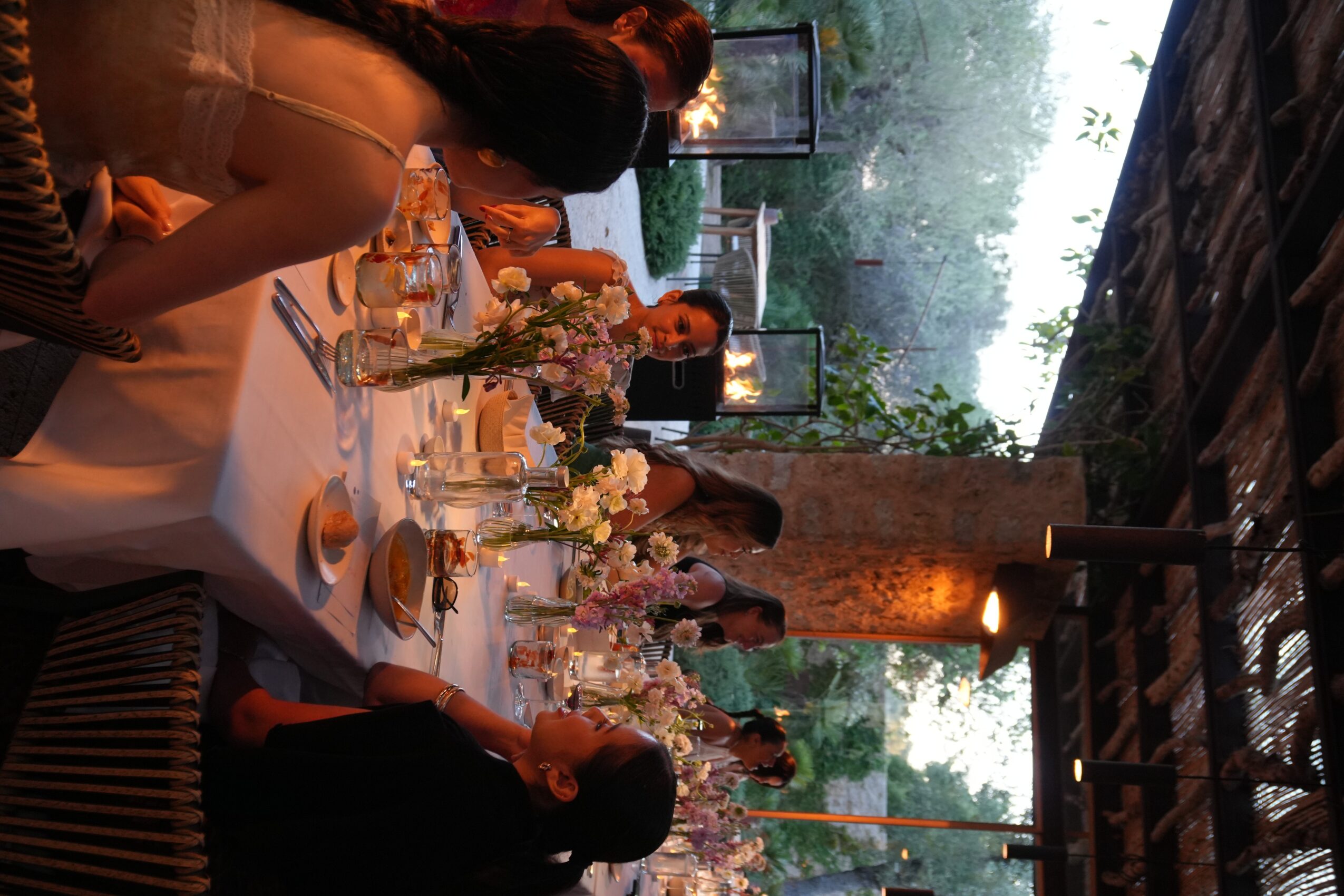 People seated at a table with flowers and food, indoors with large windows and trees outside.