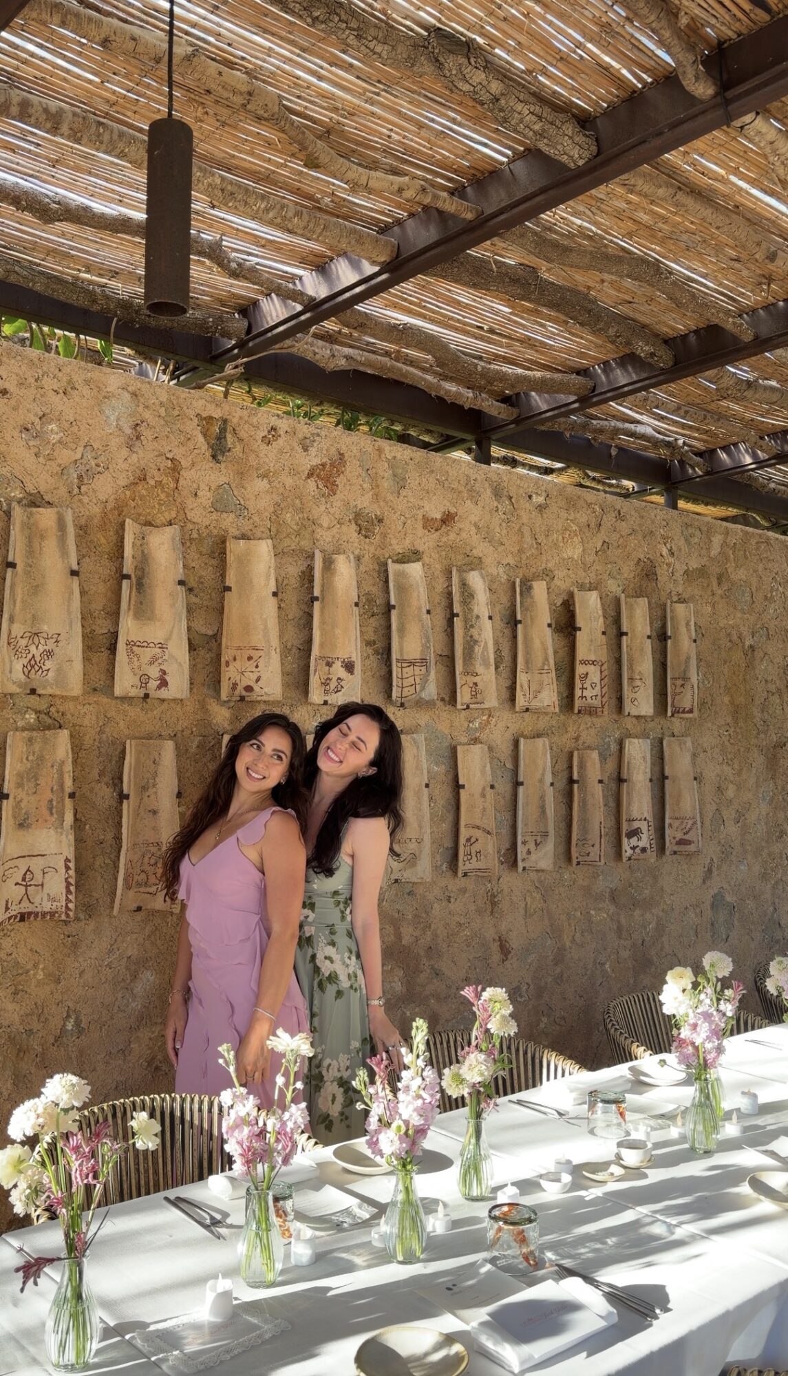 Two women standing in front of a textured wall with bamboo and wooden elements, a table with flowers and tableware in foreground.