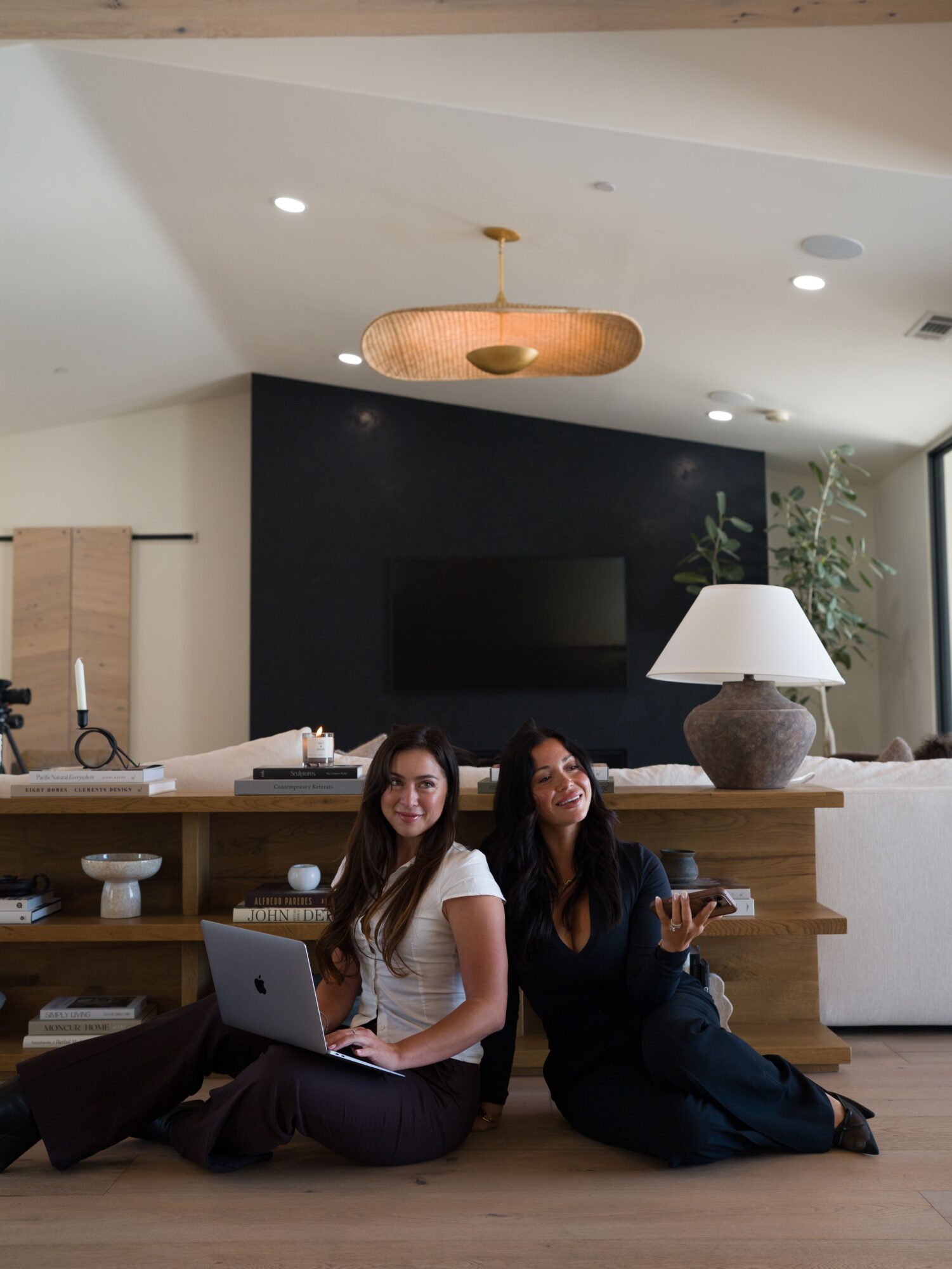 Two women sit on the floor in a modern living room with a black accent wall, TV, and a large lamp on a wooden shelf.
