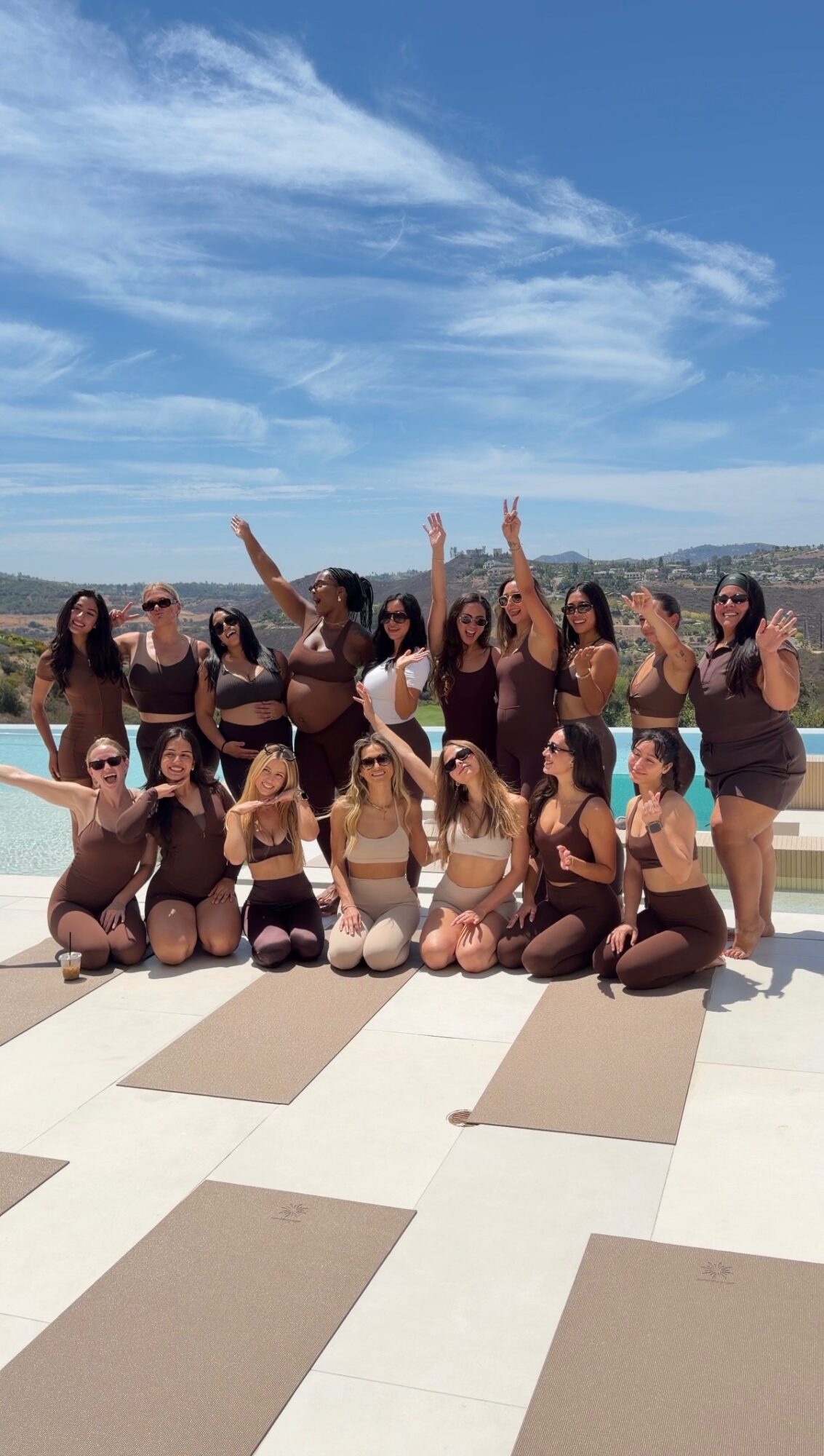 Group of women in swimsuits posing outdoors on a sunny day with a body of water and mountains in the background.