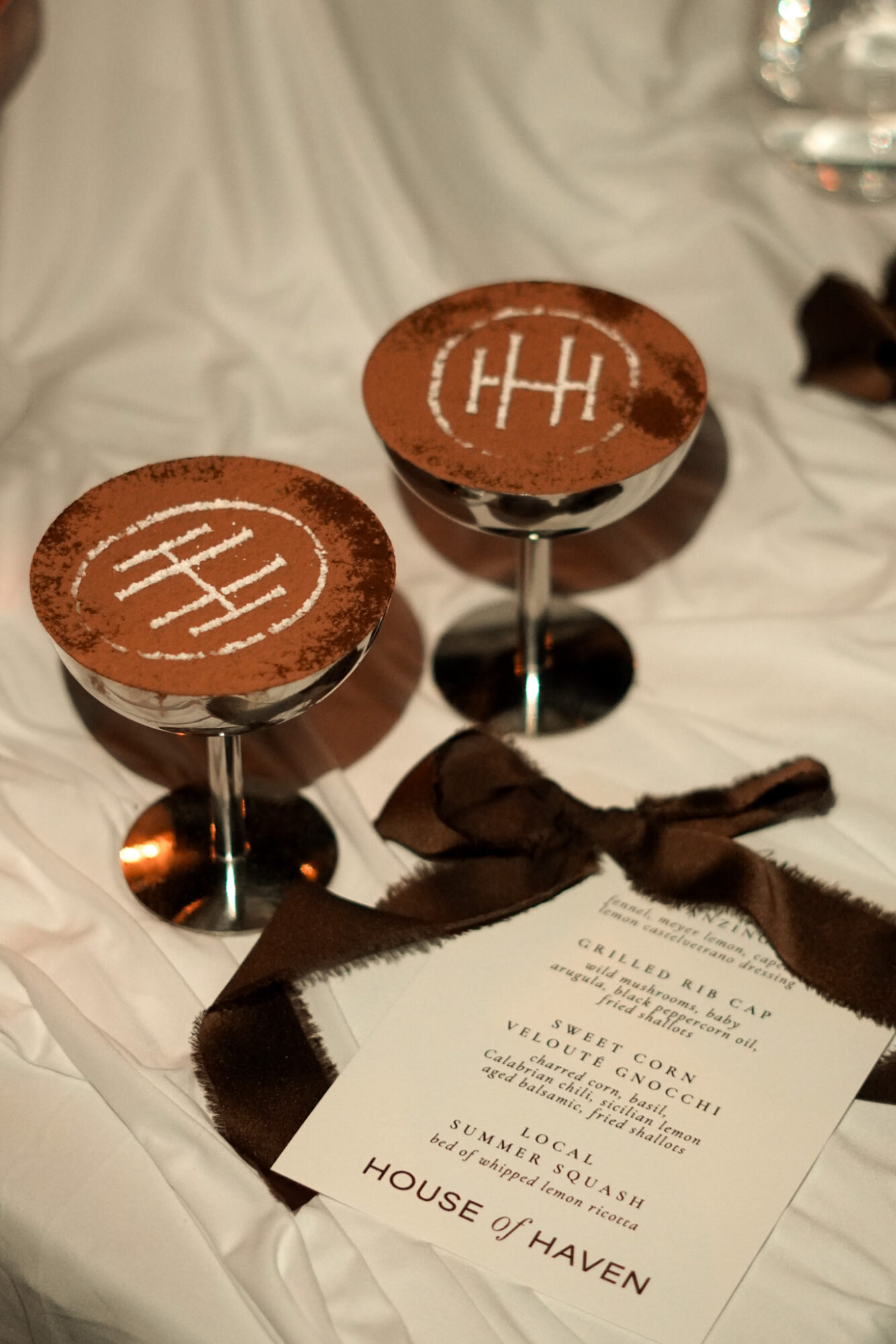 Two glasses with chocolate desserts decorated with tic-tac-toe patterns, a black ribbon, and a menu on a white tablecloth.