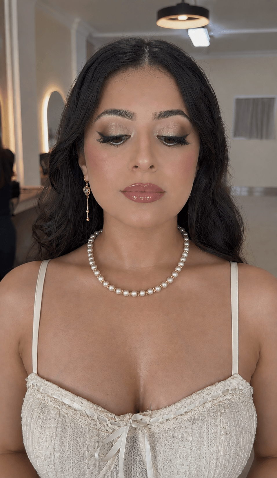 Young woman with dark hair, makeup, pearl necklace, and earrings, wearing a cream-colored top with lace details.