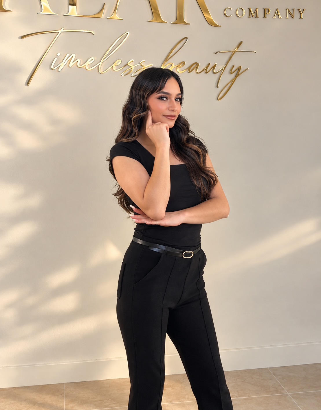 Woman with long dark hair in black outfit standing against a light background with gold text, smiling and resting her chin on her hand.